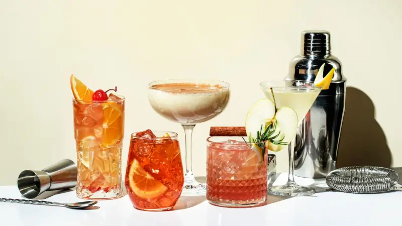 Eine Auswahl an fruchtigen Cocktails, garniert mit Orangen, Äpfeln und Kirschen, neben einem Cocktail-Shaker.
