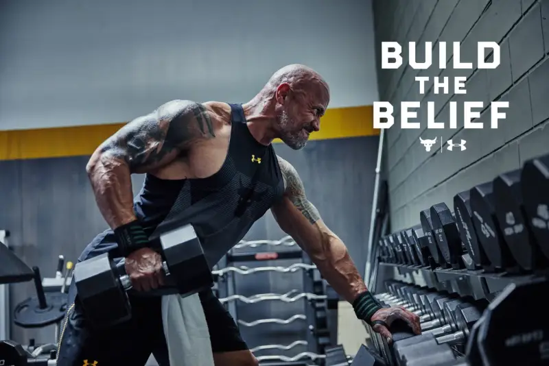 Dwayne "The Rock" Johnson w koszulce Under Armour wykonuje ćwiczenia z hantlami. W tle stojaki z ciężarami.