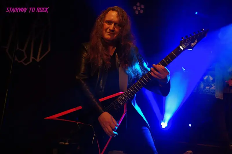 Guitarrista con pelo largo y chaqueta de cuero, tocando una guitarra eléctrica negra con detalles rojos. El escenario está iluminado con luces azules, creando un ambiente de rock.
