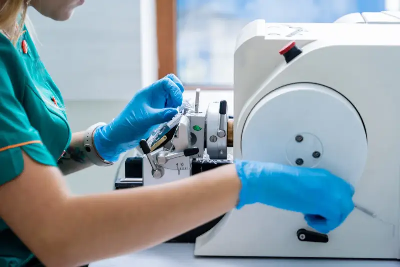 Ręce w niebieskich rękawiczkach pracują przy mikrotomie, przygotowując próbkę tkanki do badania. Może to być kluczowe dla diagnozy, czy wykryty guz jest złośliwy.
