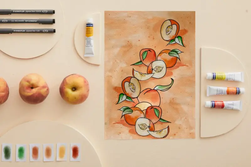 Une peinture de fruit aquarelle, avec des pêches entières et coupées, des tubes de peinture et des feutres.