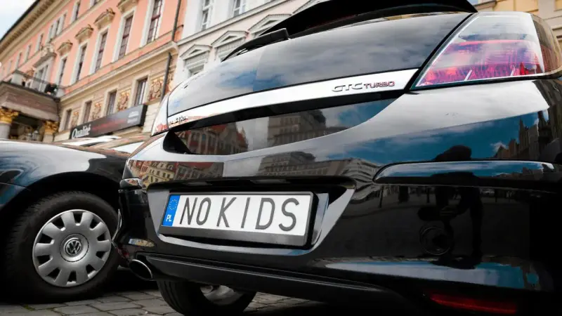 Czarne auto z indywidualnymi tablicami rejestracyjnymi "NO KIDS" na tle kamienicy.