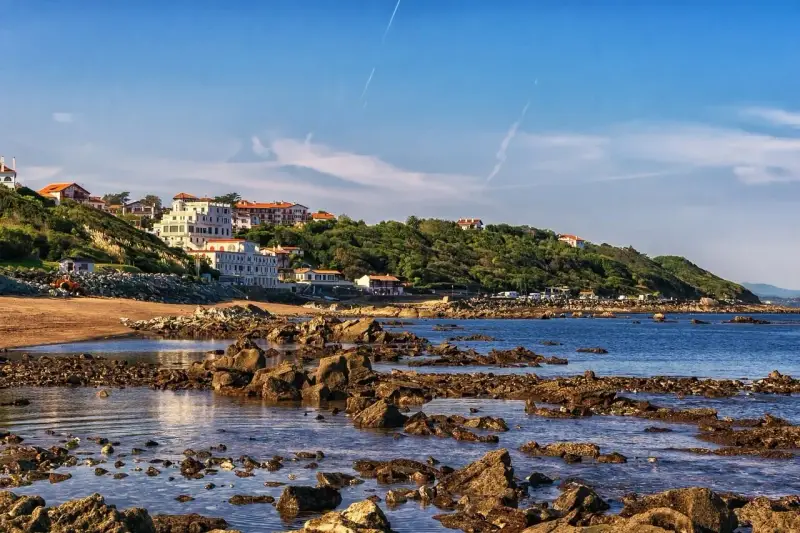 Vue côtière avec des maisons blanches sur une colline verdoyante, une plage de sable et des rochers dans l'eau. Le cadastre zone inondable est visible.
