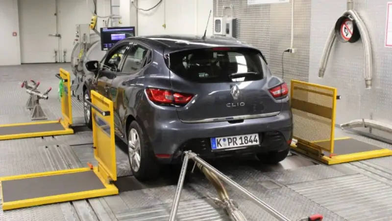 A dark grey Renault Clio is on a dynamometer in a testing facility, with exhaust pipes connected.