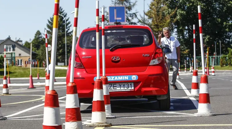 Czerwony Hyundai na placu manewrowym, z literą "L" na dachu. Instruktor pokazuje znak pokoju. Przygotowania do egzaminu na prawo jazdy b+e.
