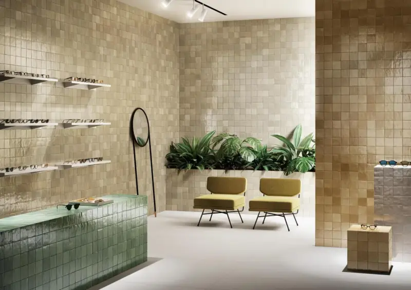 Un espace moderne avec des murs recouverts de **zellige** beige, un comptoir vert émeraude, des plantes luxuriantes et des chaises jaunes.