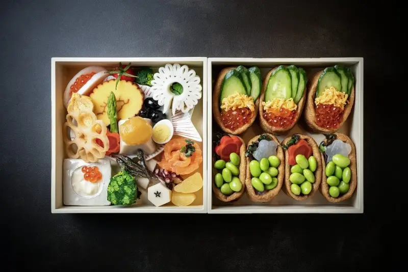 Dwa pudełka z japońskimi przysmakami: jedno z różnorodnymi składnikami, drugie z inari sushi na słodko, wypełnione edamame i ikrą.