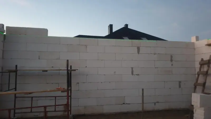 Budowa domu w toku. Ściany z bloczków betonowych czekają na tynkowanie. W tle widać dach i kominy.