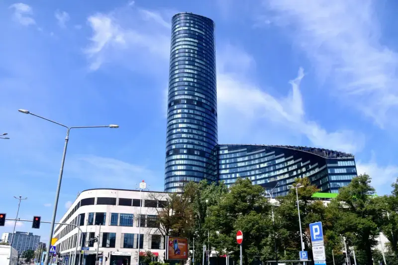 Wysoki, szklany budynek Sky Tower we Wrocławiu, z pewnością ma wiele pięter. Widać też niższy budynek i drzewa.