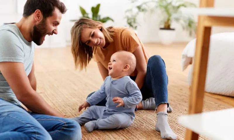 Szczęśliwa rodzina spędza czas z dzieckiem. Radosna opieka na dziecko 2 dni, pełna uśmiechów i zabawy na dywanie.