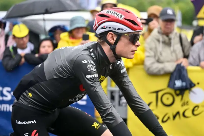 Trine Vingegaard, a cyclist in a grey and black racing suit, rides in the rain. Spectators watch from behind a yellow barrier with "Tour de France" branding.