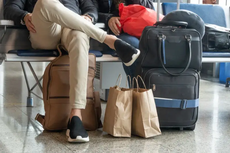 Osoby czekają na lotnisku z bagażem. Widać plecak, torby podróżne i papierowe torby na zakupy. To typowy nouvelair bagaż.
