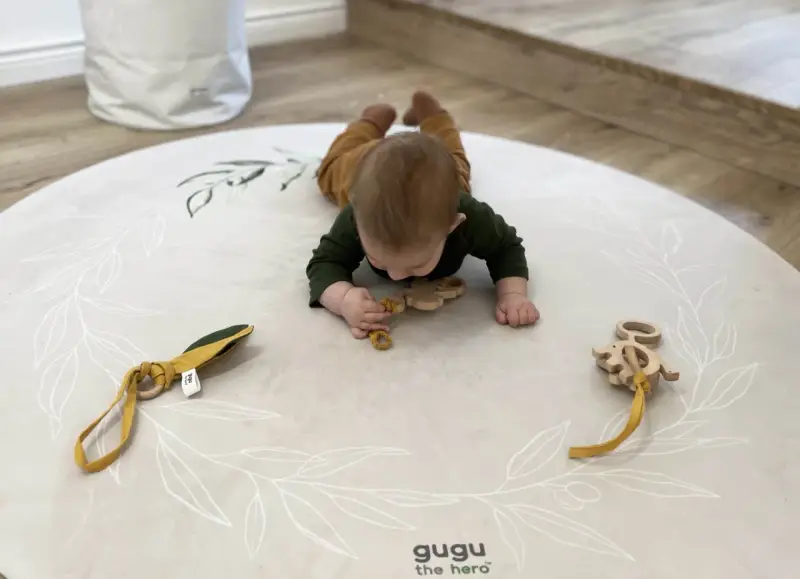 Niemowlę na brzuchu na macie z motywem roślinnym bawi się drewnianymi gryzakami.