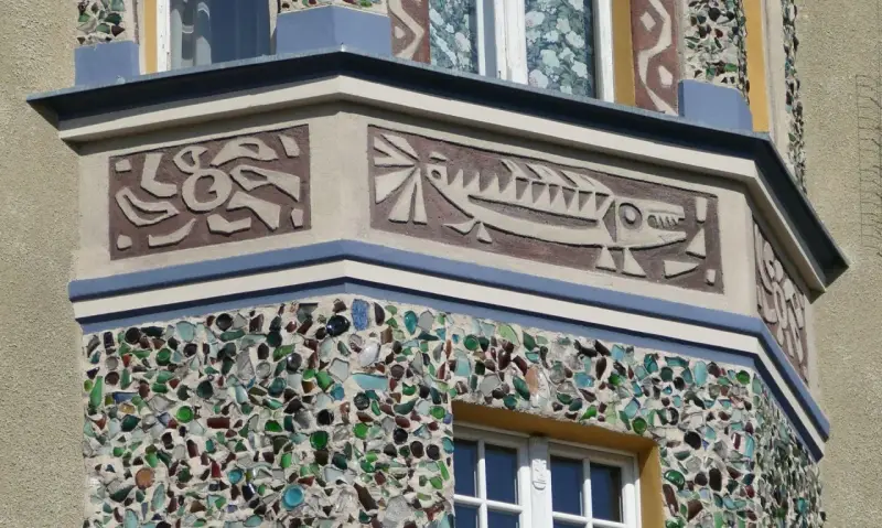Sgraffito na fasadzie budynku: stylizowana ryba i abstrakcyjne wzory. Dolna część ozdobiona mozaiką z kolorowego szkła.