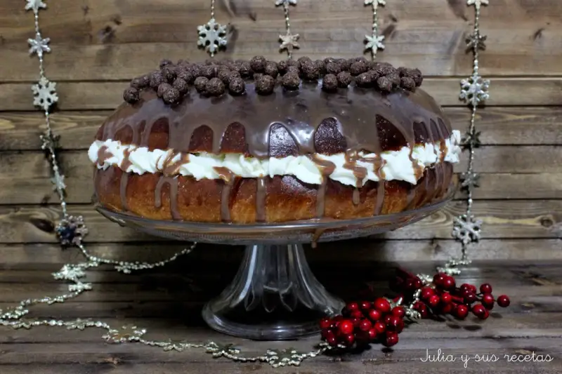 Un delicioso roscon de reyes chocolate, cubierto de glaseado y bolitas de chocolate, con crema y decoraciones navideñas.