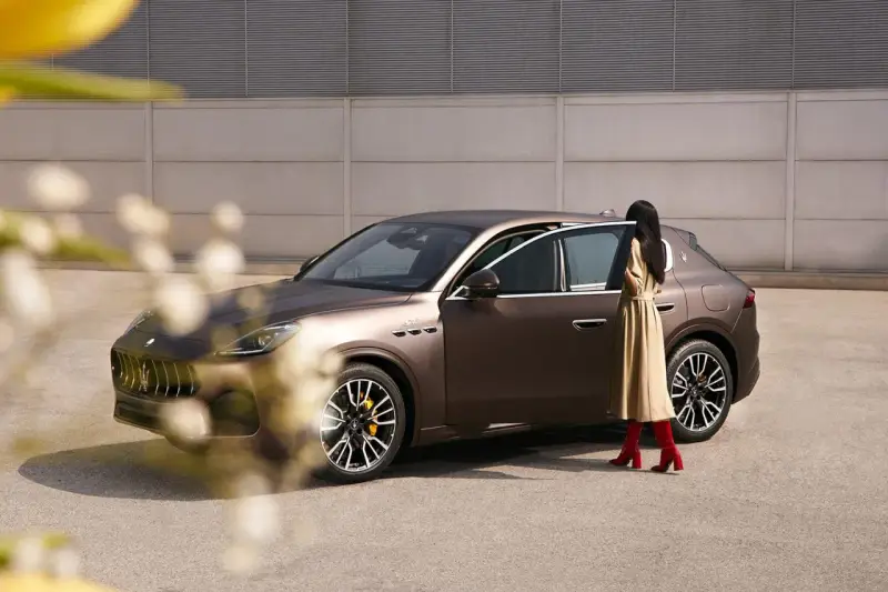 Elegancka kobieta w beżowej sukience i czerwonych kozakach zbliża się do brązowego Maserati SUV.