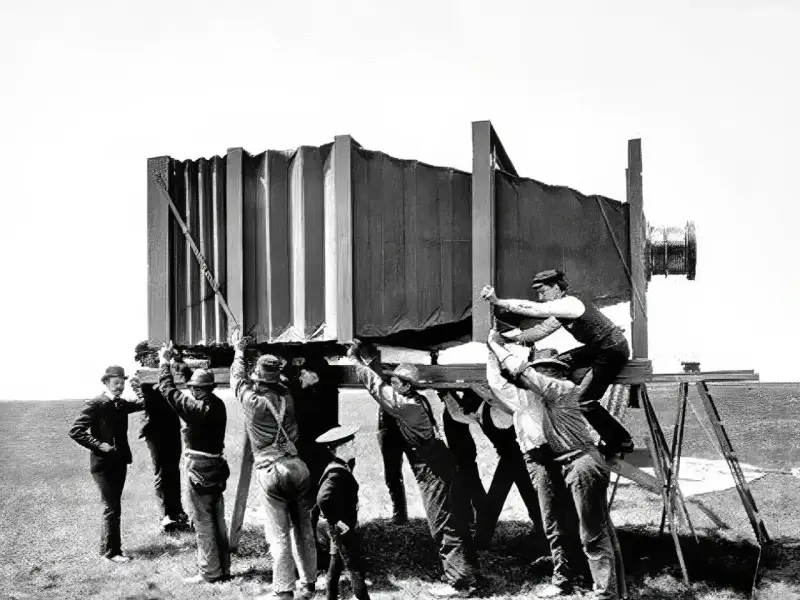 Grupa mężczyzn w historycznych strojach ustawia gigantyczny aparat fotograficzny. Wyobraź sobie, kiedy wynaleziono aparat fotograficzny i jak wielkie musiały być pierwsze urządzenia!