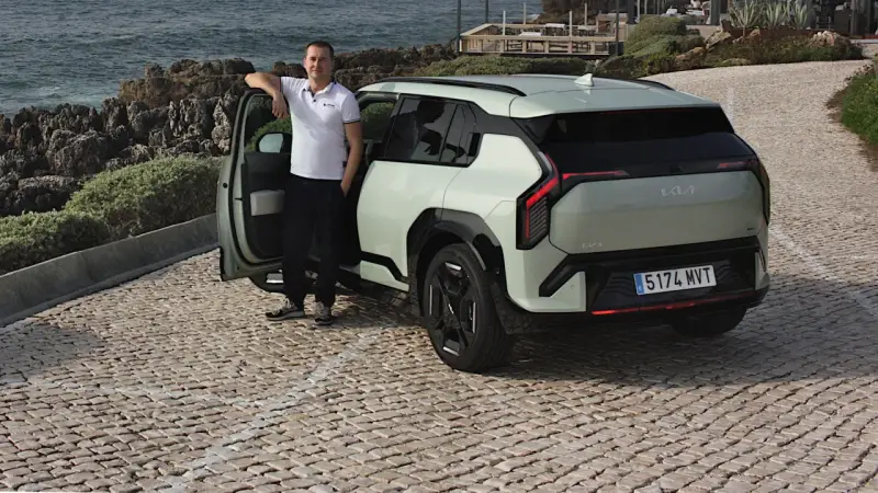 Mężczyzna opiera się o drzwi nowego, jasnoszarego Kia EV3, zaparkowanego na brukowanej drodze nad morzem.