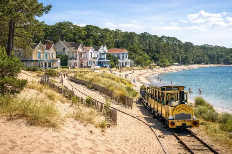 Petit train jaune longeant une plage de sable bordée de maisons colorées et d'une forêt.