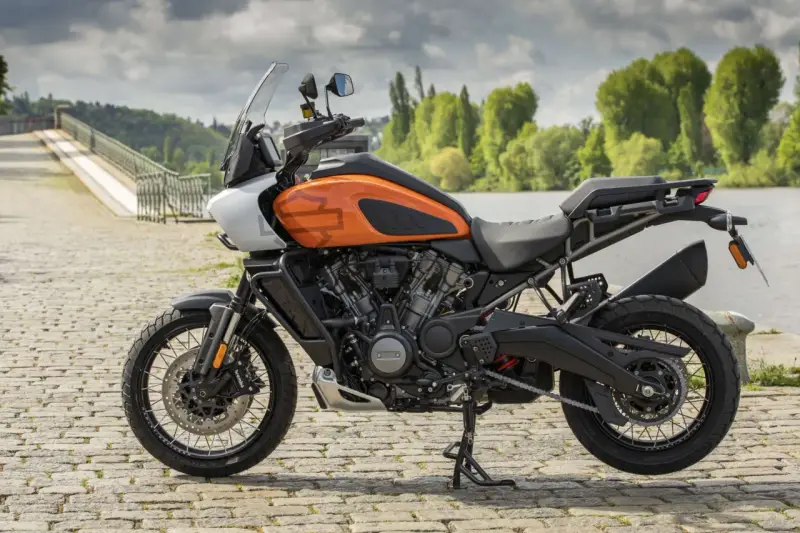 Pomarańczowy Harley-Davidson Pan America gotowy na przygodę, z mostem i zielenią w tle.