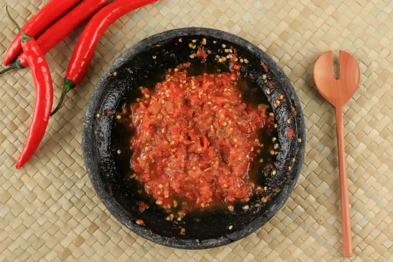 Świeżo przygotowany sos sambal w kamiennym moździerzu, obok czerwone papryczki chili i drewniana łyżka.