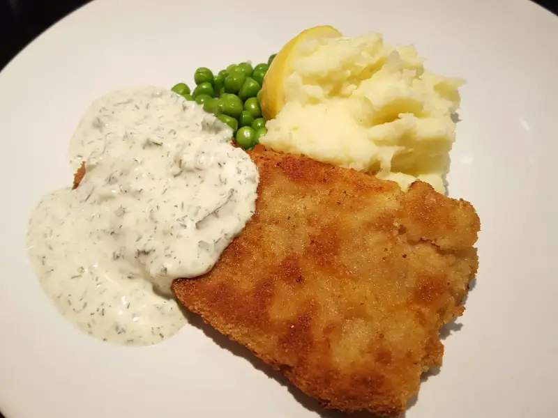 Panerad fisk med krämig kall sås till, serverad med potatismos, gröna ärtor och en citronklyfta.