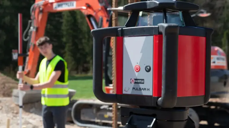 Niwelator laserowy Pulsar H na budowie. W tle pracownik w kamizelce odblaskowej i koparka.
