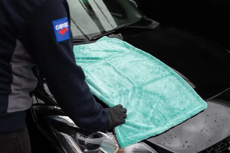 Person i handskar lägger en turkos mikrofiberduk på en svart bilhuv. Duken är från the rag company.