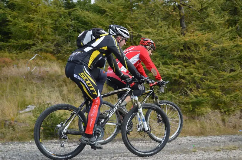 Dwóch kolarzy górskich w kaskach i strojach sportowych jedzie po szutrowej drodze. Jeden z nich, z tyłu, ma na nogawkach napis "Scott". Słychać, jak rower skrzypi podczas pedałowania.