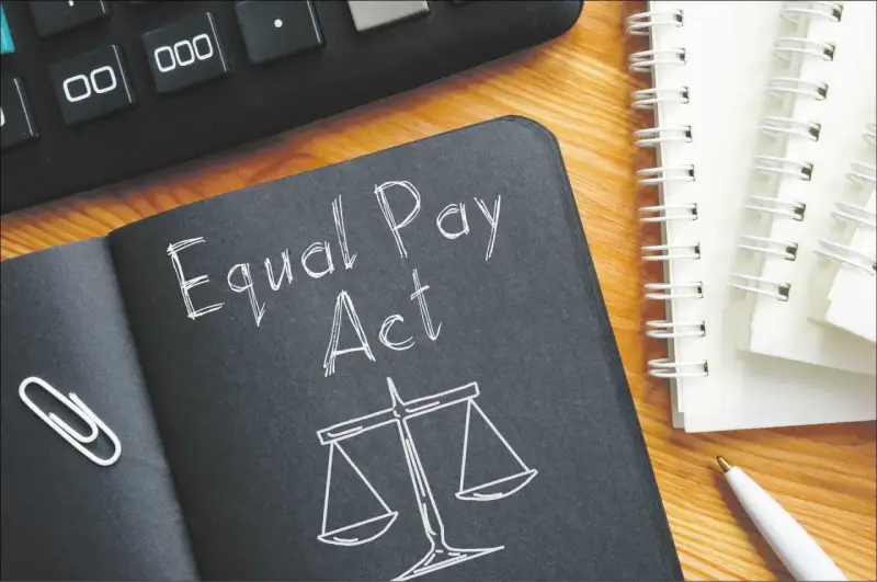 Notatnik z napisem "Equal Pay Act" i wagą sprawiedliwości symbolizuje jawność wynagrodzeń. Obok kalkulator i długopis.