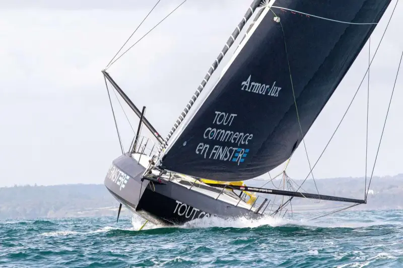 Le voilier IMOCA de Jean Le Cam fend les vagues, sa grand-voile noire ornée du slogan "Tout commence en Finistère" gonflée par le vent.