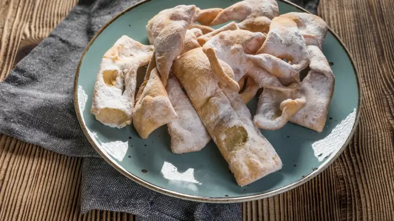 Chrupiące faworki, przygotowane według przepisu babci, posypane cukrem pudrem, leżą na talerzu.