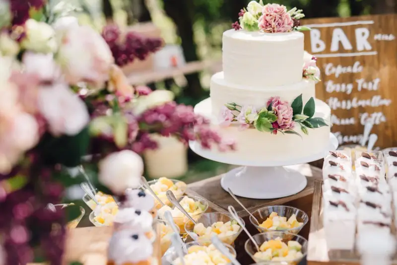 Elegancki tort weselny boho ozdobiony kwiatami, obok deserki i owoce.