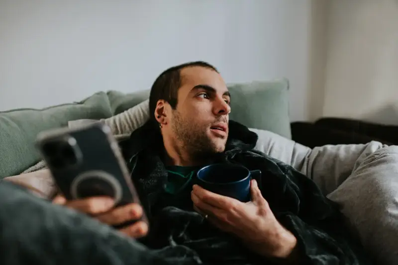 Mann liegt auf Sofa, hält Tasse und Handy. Mögliche Bewegungsmangel Symptome wie Müdigkeit und Lethargie werden angedeutet.