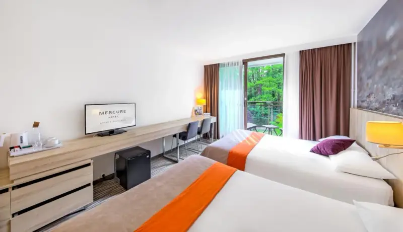 Pokój hotelowy z dwoma łóżkami, biurkiem i balkonem z widokiem na zieleń. Hotel Posejdon Gdańsk.