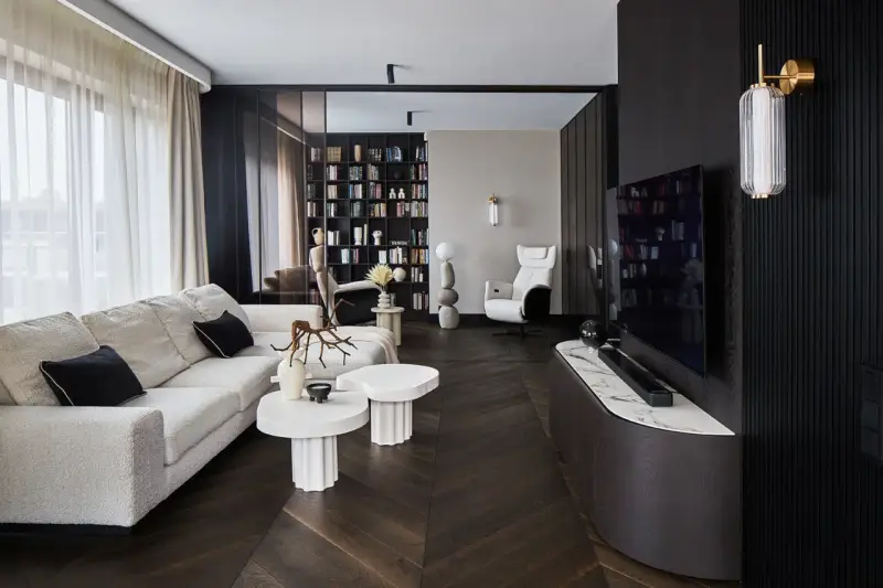 Nowoczesny salon z wygodną sofą, stolikami i regałem pełnym książek. Dominują ciemne, grafitowe akcenty, tworząc elegancką atmosferę.