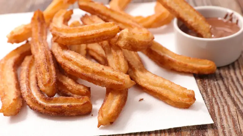Goldbraune Churros, frisch frittiert, mit Zucker bestreut. Ein Churro wird in eine Schüssel mit Schokoladensauce getunkt. Ein echtes Churros Rezept Original.