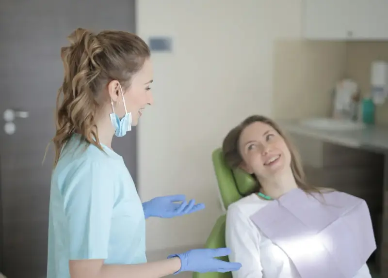 Uśmiechnięta pacjentka na fotelu stomatologicznym słucha dentystki. To obrazek z gabinetu, gdzie odbywają się studia stomatologiczne.