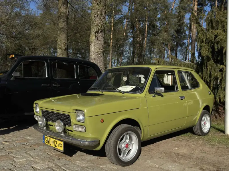 Zielony fiat 127 z lat 70. stoi obok czarnej taksówki na tle drzew.