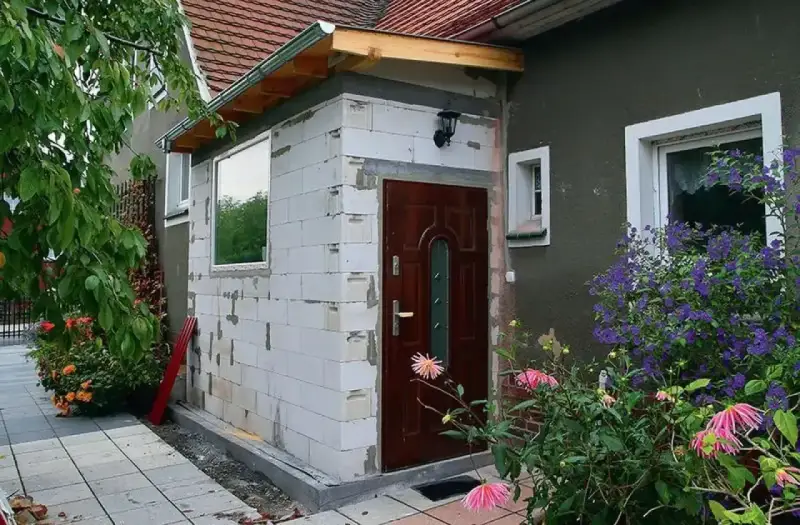 Budowa nowego przedsionka do domu bez pozwolenia. Białe pustaki, brązowe drzwi i okno czekają na wykończenie.
