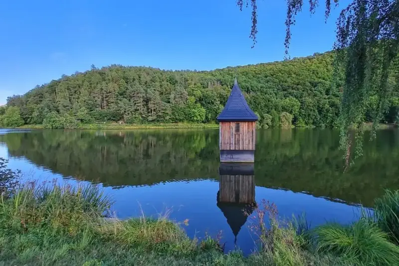 Holzhaus mit spitzem Dach, wie eine kleine Kirche im See, spiegelt sich im ruhigen Wasser.