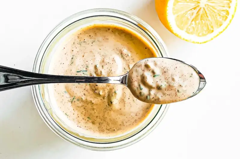 En sked krämig remouladsås med örter lyfts ur en burk. Bredvid ligger en halv citron. Perfekt för ditt remouladsås recept!