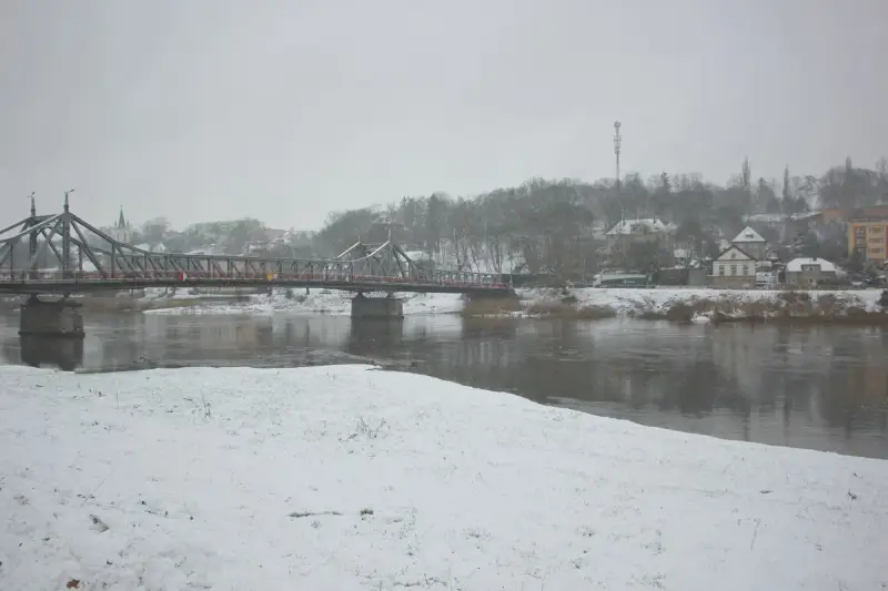 Most na rzece w zimowy dzień, śnieg pada. W tle zabudowania i drzewa. To piękna dx rejestracja krajobrazu.