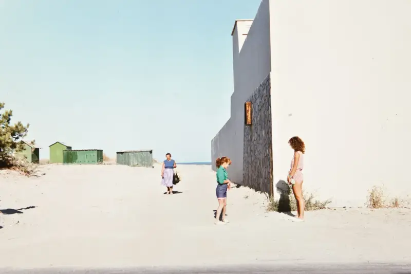 Luigi Ghirri foto: una donna cammina sulla spiaggia, due bambine giocano vicino a un edificio bianco.