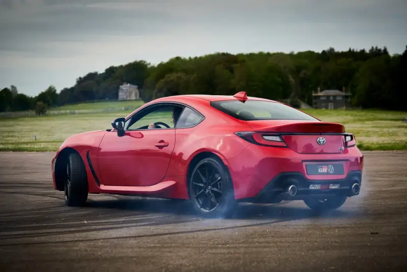 Czerwona Toyota 86 GR w poślizgu, z opon dymiących na asfalcie. W tle zielone pola i drzewa.