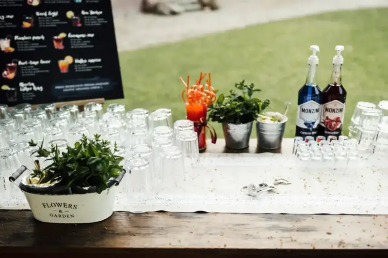 Stół z kieliszkami, butelkami syropów i miętą, gotowy na przygotowanie drinków na wesele. Przepisy na drinki są widoczne na tablicy w tle.