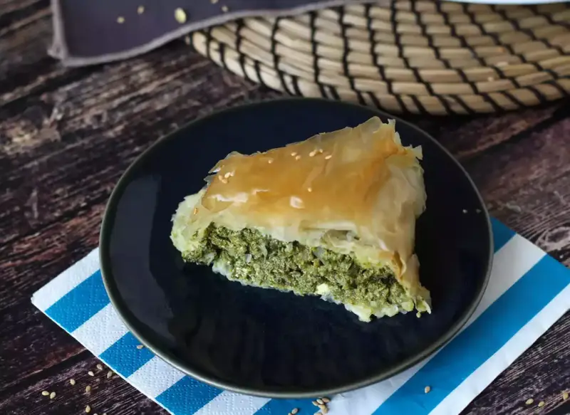 Kawałek spanakopity z chrupiącym ciastem filo i zielonym nadzieniem ze szpinaku i sera feta, posypany sezamem.
