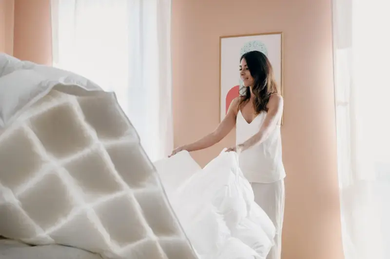Donna sorride mentre sistema un piumone bianco, mostrando come piegare un piumone per un letto ordinato.