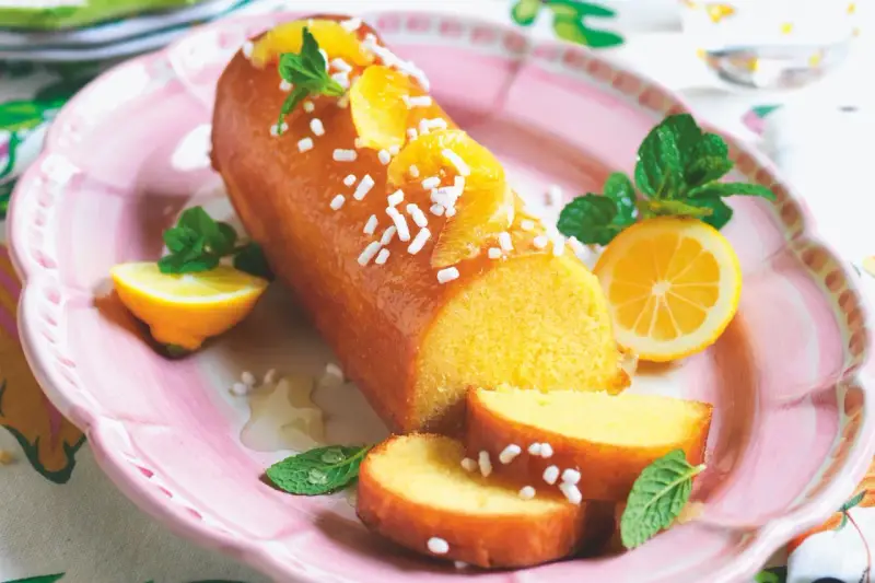 Deliziosa torta al limone glassata, decorata con fette d'arancia, zuccherini e menta. Servita su un piatto rosa con spicchi di limone.