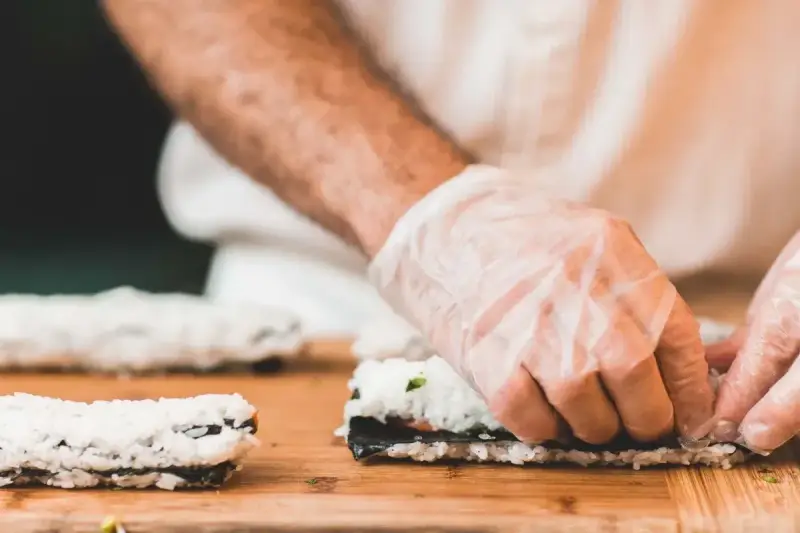Ręce w rękawiczkach zwijają sushi. Na desce leżą gotowe rolki i składniki do kolejnej. Idealna zalewa do sushi podkreśli smak.